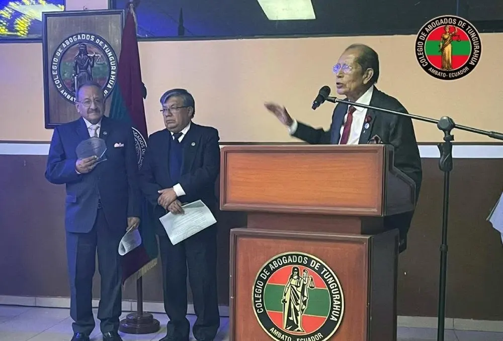Orador hablando en el podio con el emblema del Colegio de Abogados de Tungurahua, acompañado por dos abogados